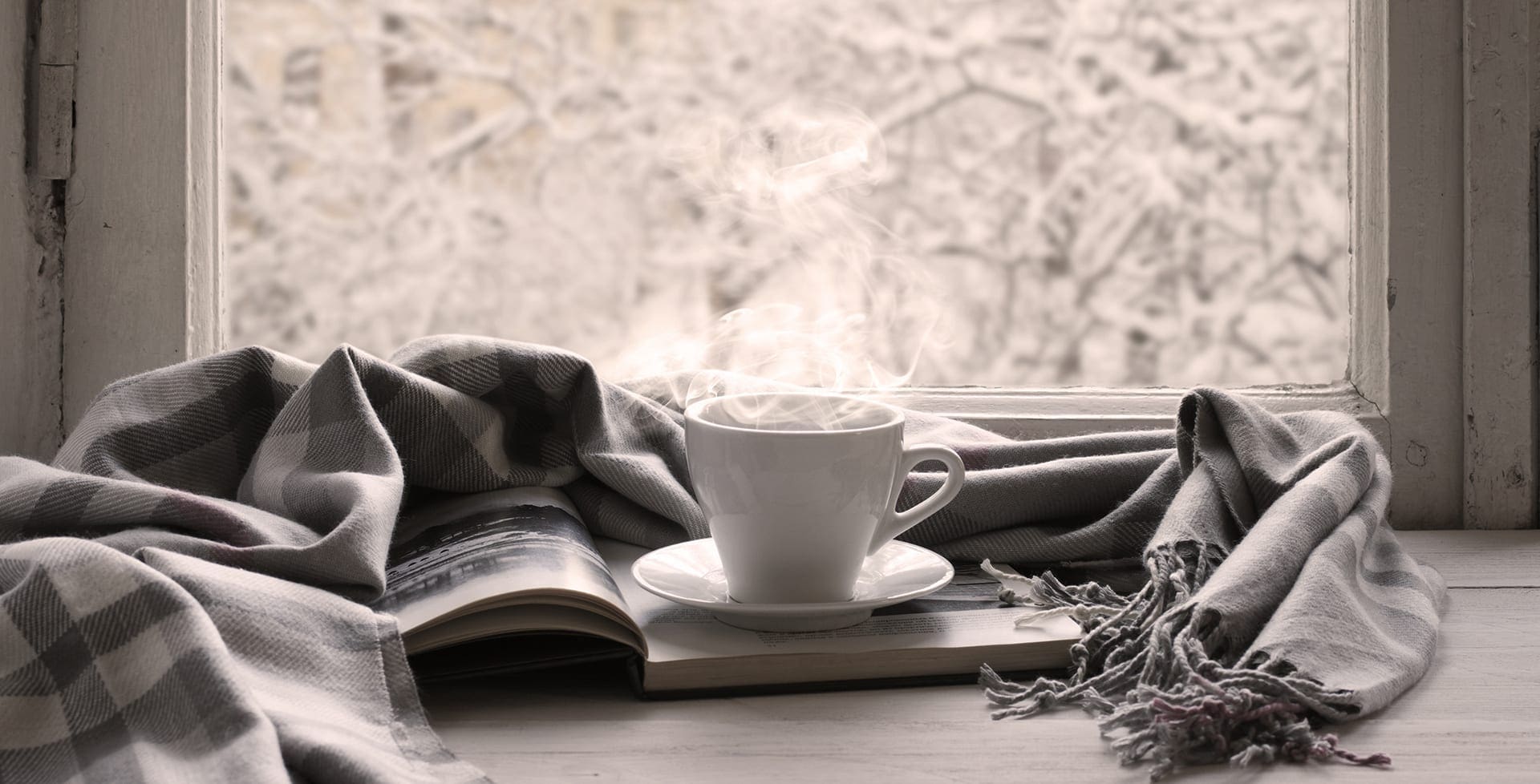 Cozy winter still life: cup of hot coffee and opened book with warm plaid on vintage windowsill against snow landscape from outside.